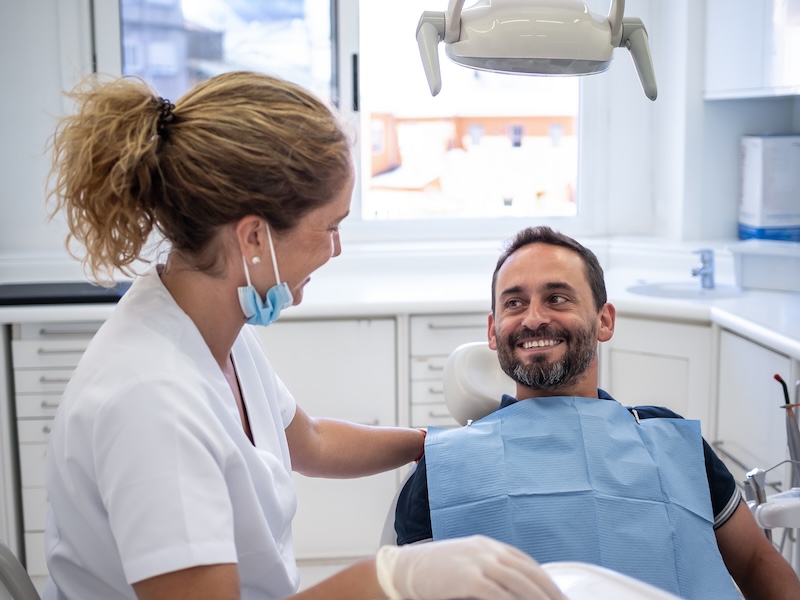 A patient sits in a dentist's office alongside his dentist, wearing a dental bib. The dentist is explaining what wisdom teeth removal near Hampton, MD, is and what he can expect from the procedure.